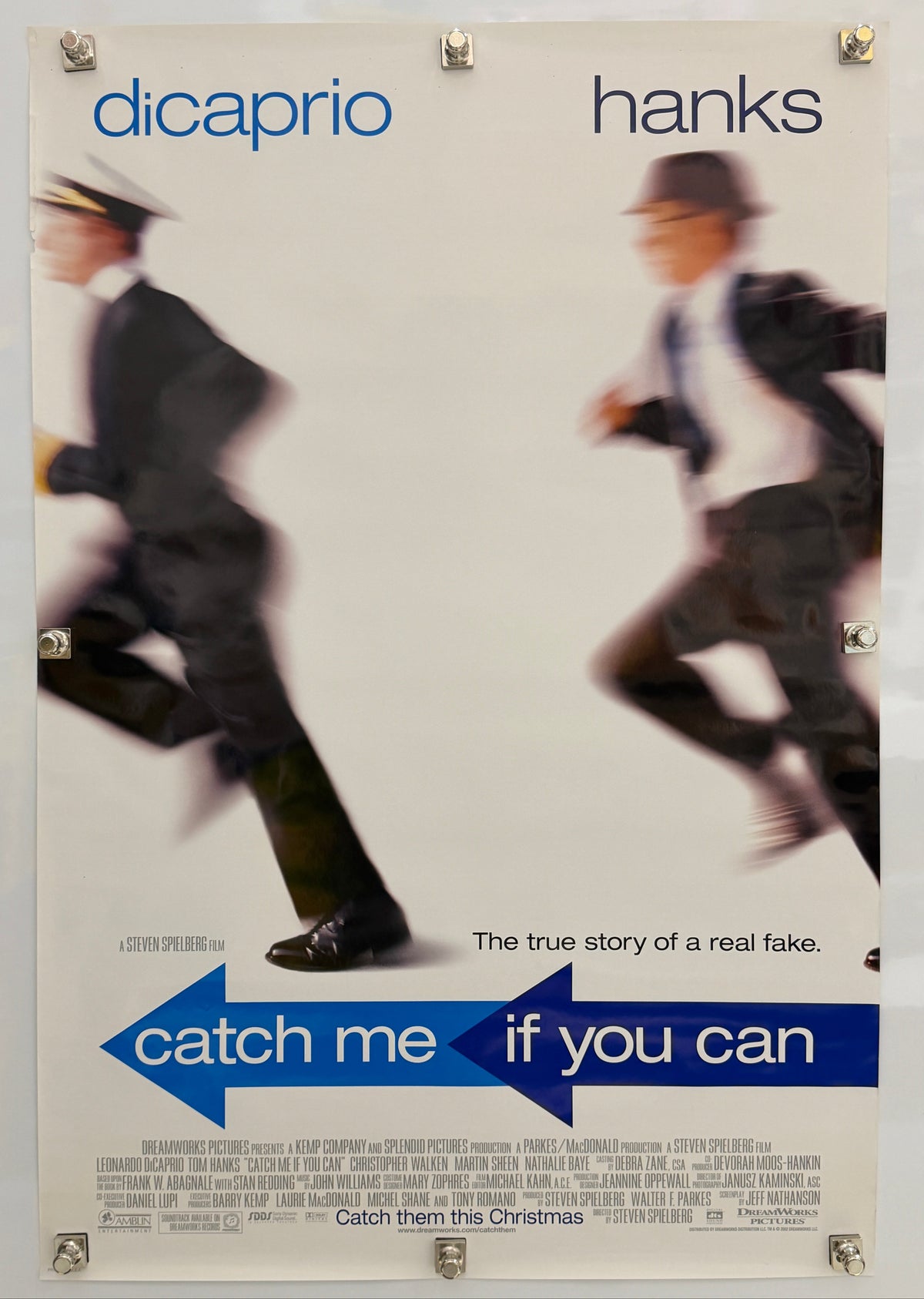 A vintage movie poster for 'Catch Me If You Can' featuring blurred figures of two men in suits, with blue title text and the names 'hanks' and 'dicaprio' printed on a light background.