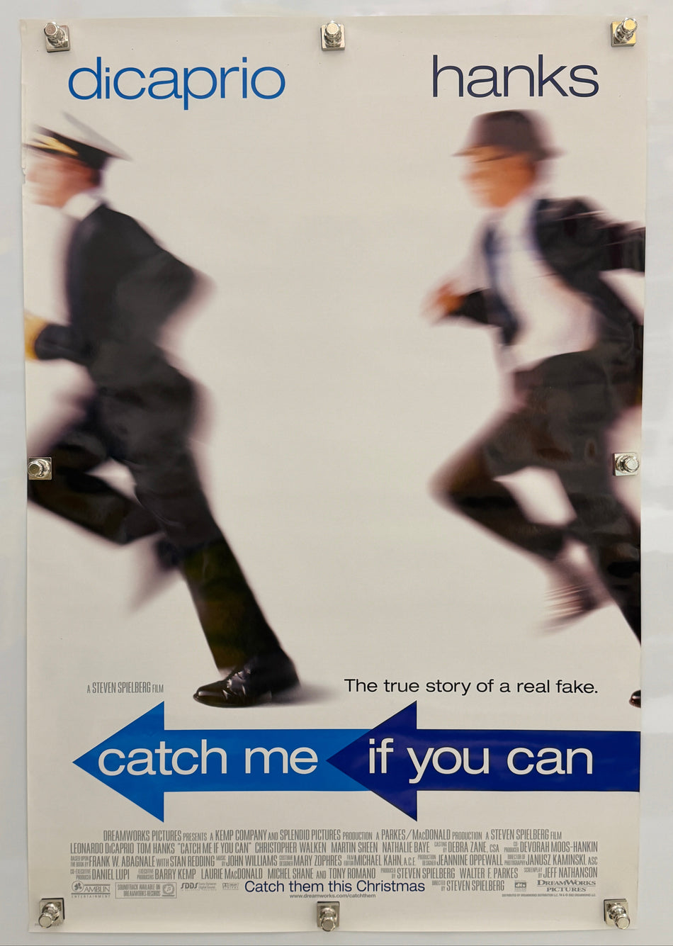A vintage movie poster for 'Catch Me If You Can' featuring blurred figures of two men in suits, with blue title text and the names 'hanks' and 'dicaprio' printed on a light background.
