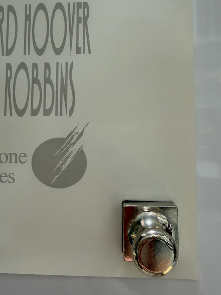A close-up of a white printed card or label with partial gray text reading 'HOOVER ROBBINS' and a circular logo mark, secured with a shiny silver metal clip in the upper right corner.