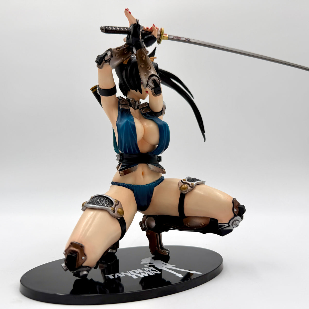 A collectible anime figurine of a female samurai warrior in a dynamic kneeling pose, holding a katana overhead. The figure has black hair, pale skin, a blue bikini-style outfit, and brown and black armor details, standing on a black oval base with 'TANKER TANK' printed on it.