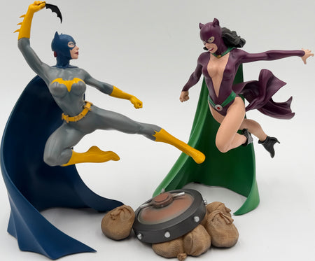 A collectible two-figure statue of Batman and Catwoman in a dynamic action pose, with Batman in gray and yellow and Catwoman in purple and green, standing on a light gray studio background.