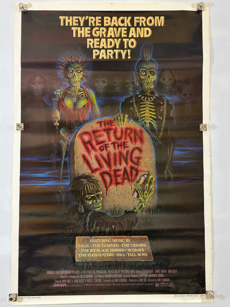A vintage horror movie poster for 'The Return of the Living Dead' taped to a white wall. The poster features zombie and skeleton characters with bold yellow and red text, and is secured with tape at the corners.
